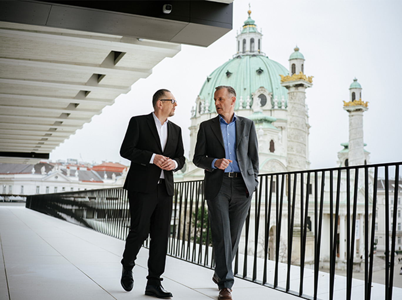 Der Generaldirektor Mag. Dr. Martin Krajcsir und der Generaldirektor-Stellvertreter DI Peter Weinelt laufen über die Terrasse des Wien Museums. Sie befinden sich im Gespräch miteinander und im Hintergrund ist die Karlskirche zu sehen.