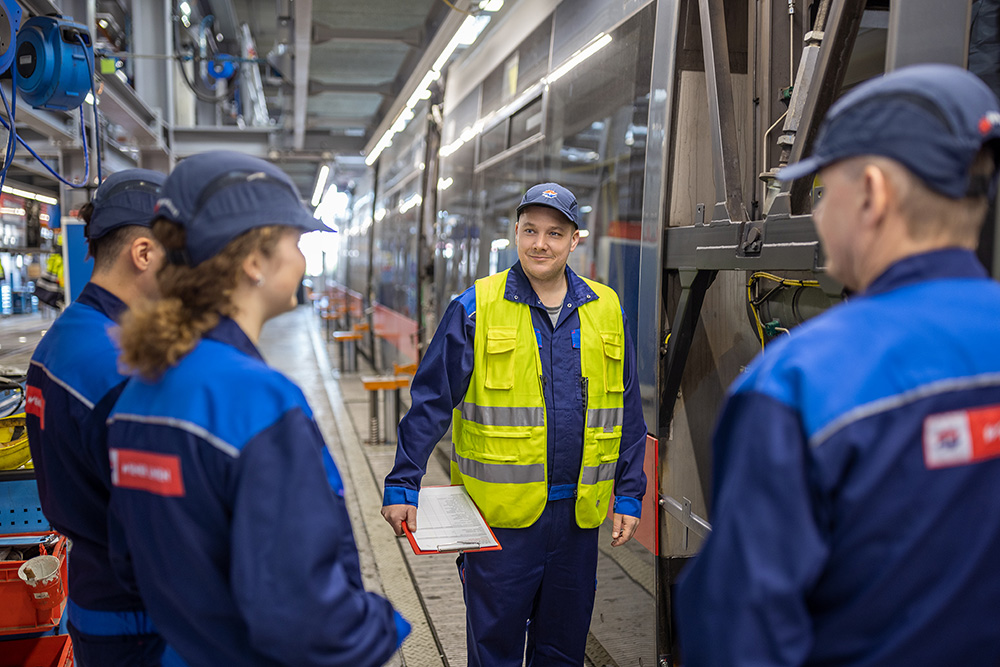 A male Wiener Netze employee is standing in a circle with three trainees and is smiling while having a conversation with them. In the background, we see Wiener Linien’s main workshop. C: Severin Wurnig