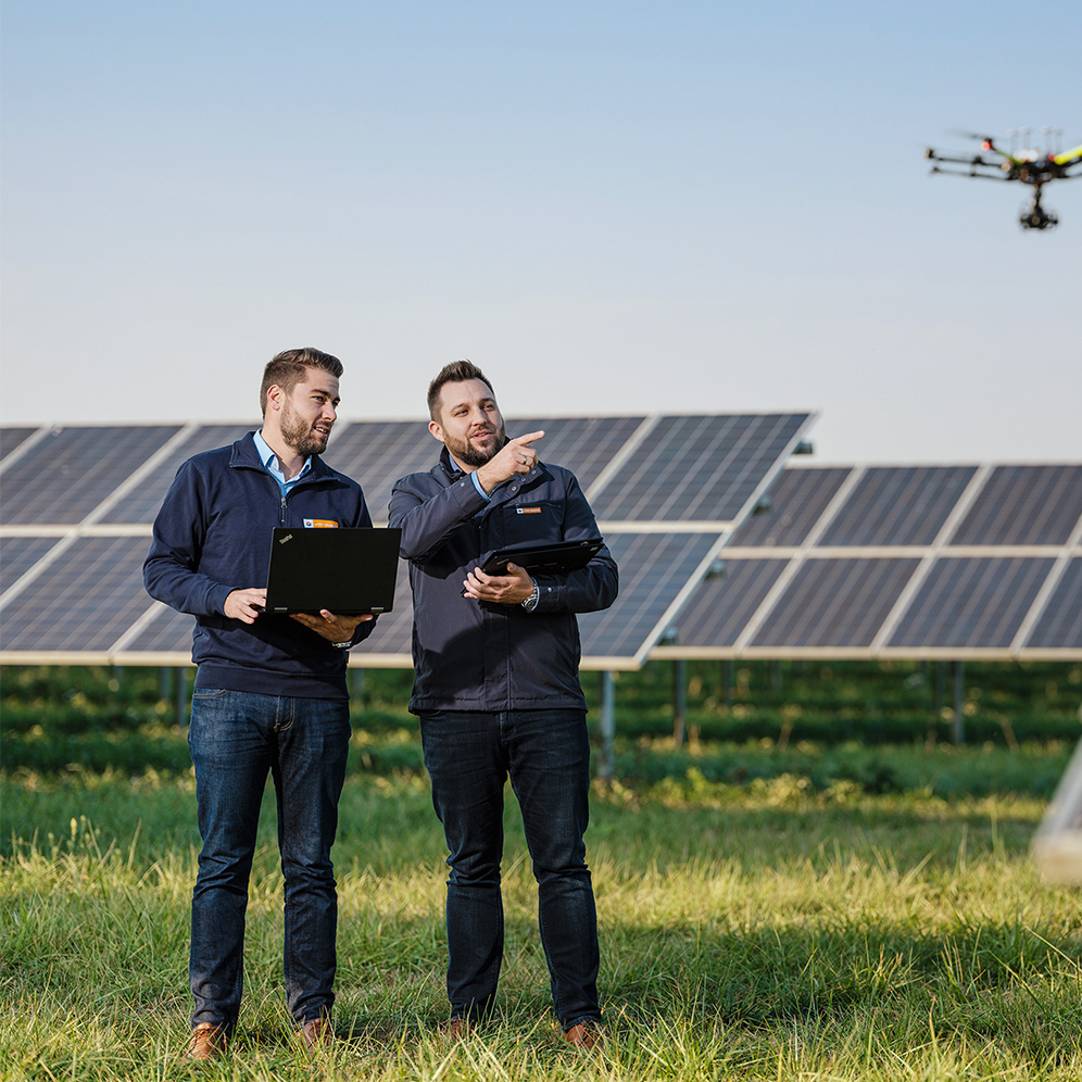 Zwei männliche Mitarbeiter stehen auf einer Rasenfläche vor einer Photovoltaikanlage. Einer der beiden zeigt auf etwas, das nicht im Bild ist. C: Wien Energie/Ehm