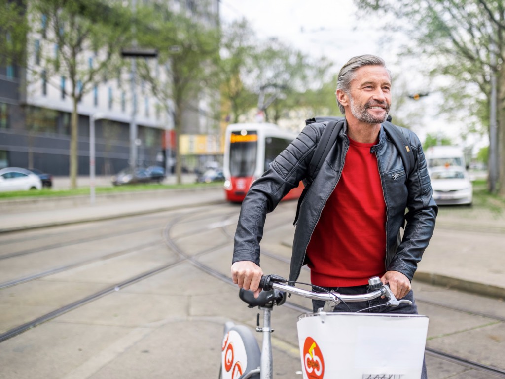 Ein Mann in schwarzer Lederjacke schiebt lachend ein Leihfahrrad durch Wien. Eine Bim fährt im Hintergrund. Foto: Wiener Stadtwerke/Severin Wurnig
