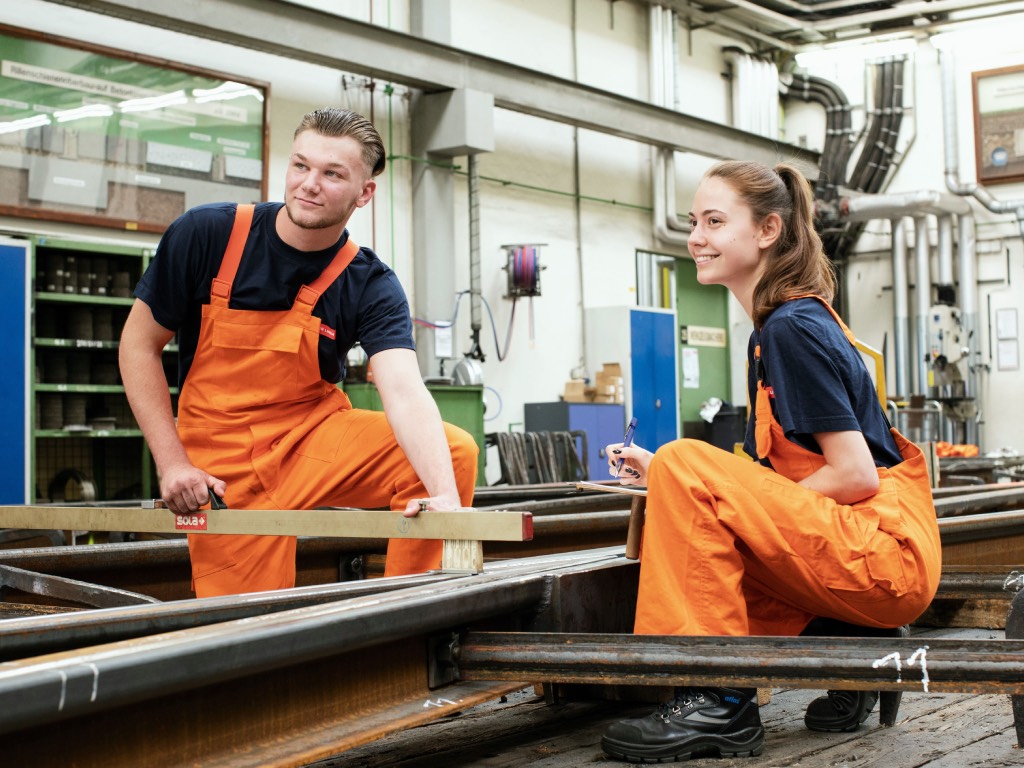 Zwei junge Mitarbeitende mit orangen Arbeitsoveralls arbeiten zusammen in einer Werkstatt. Foto: Wiener Stadtwerke/Pauty