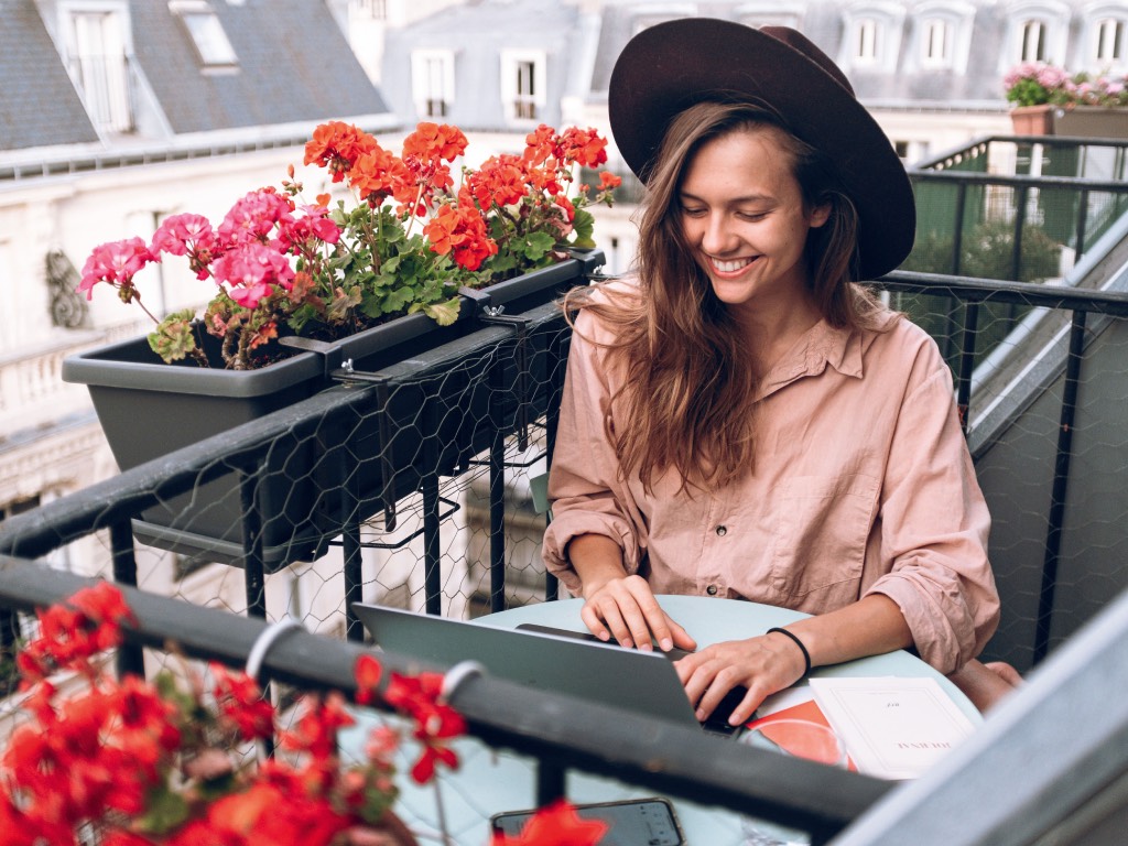 Eine junge Frau mit Hut sitzt auf ihrem kleinen Balkon und arbeitet lachend an ihrem Laptop. Foto: Pexels/Daria Shevtsova